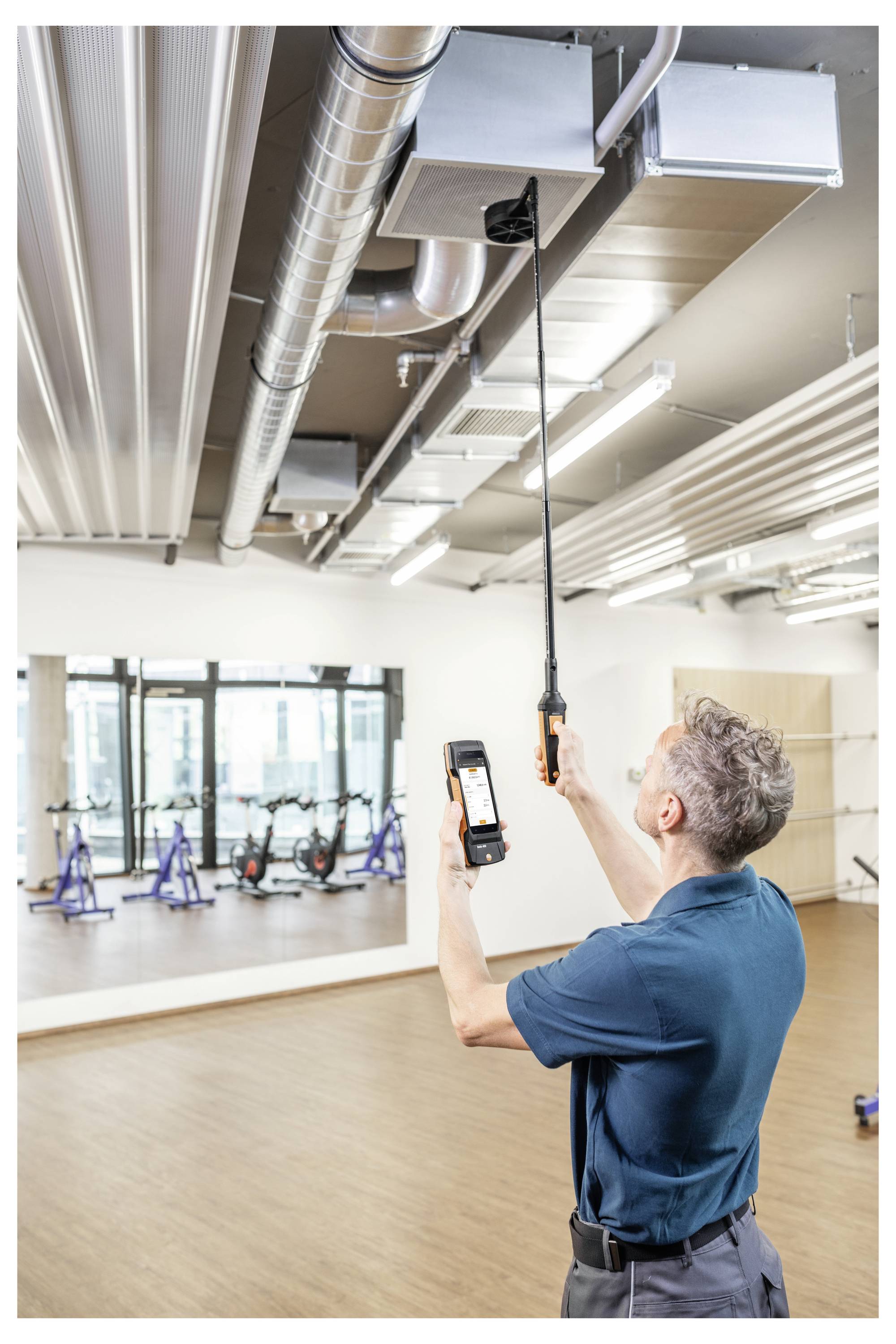Osoba w niebieskiej koszuli sprawdza kanały wentylacyjne w studio fitness, używając urządzenia ręcznego i smartfona do pomiarów.
