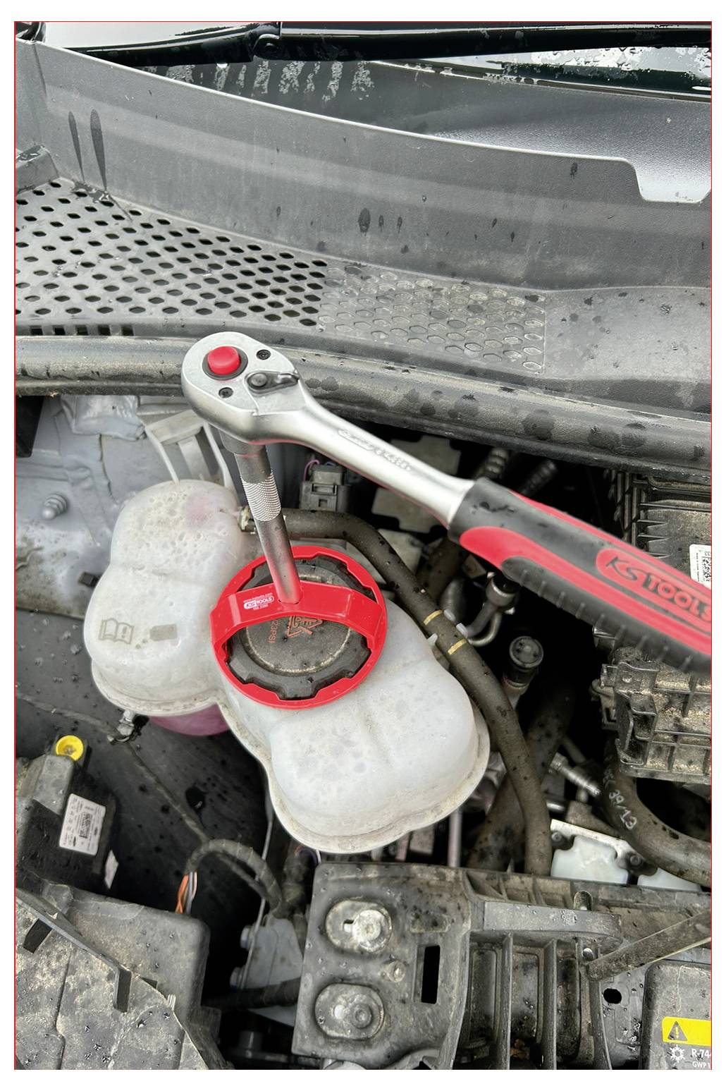 Silnik samochodowy z kluczem przyczepionym do plastikowej nakrętki zbiornika, prawdopodobnie ilustrujący sposób jej otwarcia. Narzędzie jest czerwone i metaliczne.