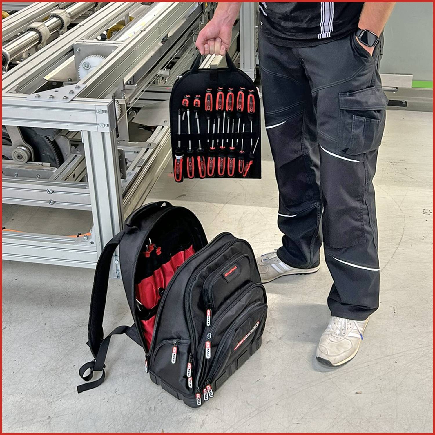 Osoba stoi przy sprzęcie przemysłowym, trzymając organizery narzędziowe z czerwonymi uchwytami. Plecak z narzędziami leży na podłodze obok niej.