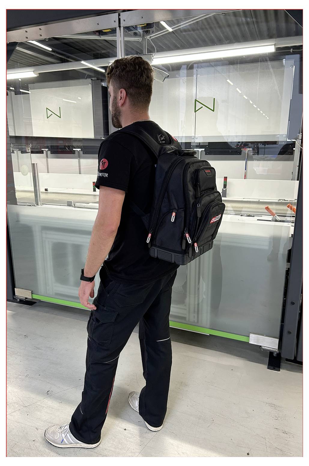 Osoba ubrana w czarny plecak i czarne ubranie stoi w nowoczesnym wnętrzu przemysłowym, zwrócona w stronę dużej maszyny z błyszczącymi, refleksyjnymi powierzchniami.