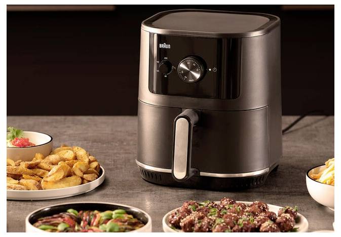 Blat kuchenny z czarnym frytkownicą powietrzną, wokół której rozłożone są talerze z frytkami, skrzydełkami z kurczaka i warzywami, sugerujący scenę przygotowywania posiłku.