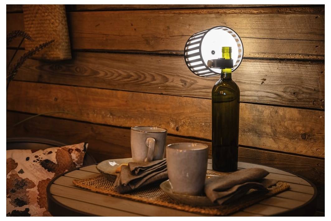 Rustykalny nakrycie stołu z dwoma kubkami, serwetkami i brązową butelką, w której umieszczono zapalony lampion, na tle drewnianej ściany, tworząc przytulną atmosferę.