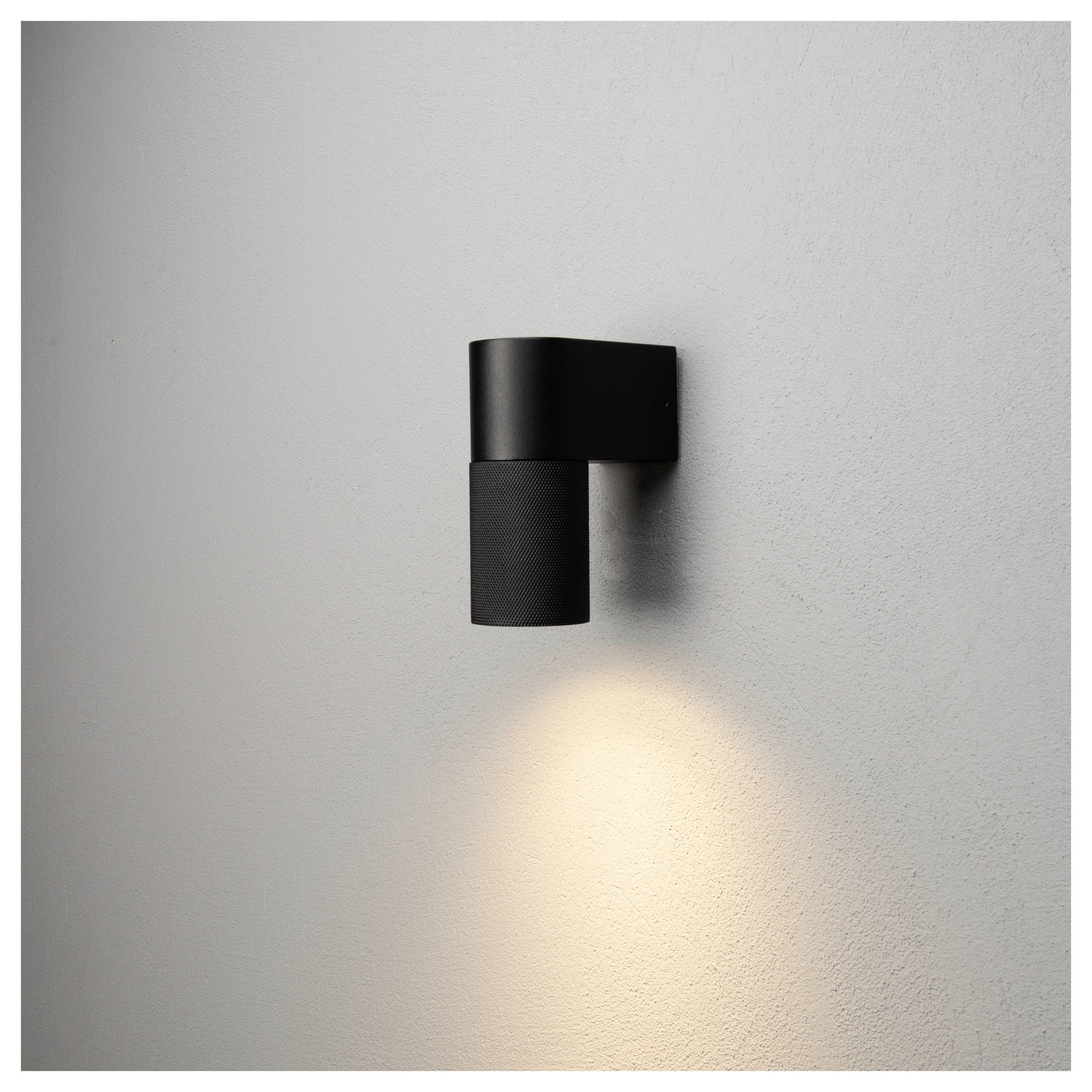 Smukła, nowoczesna czarna lampa ścienna o minimalistycznym designie rzuca ciepłe, skierowane w dół światło na gładką białą ścianę.