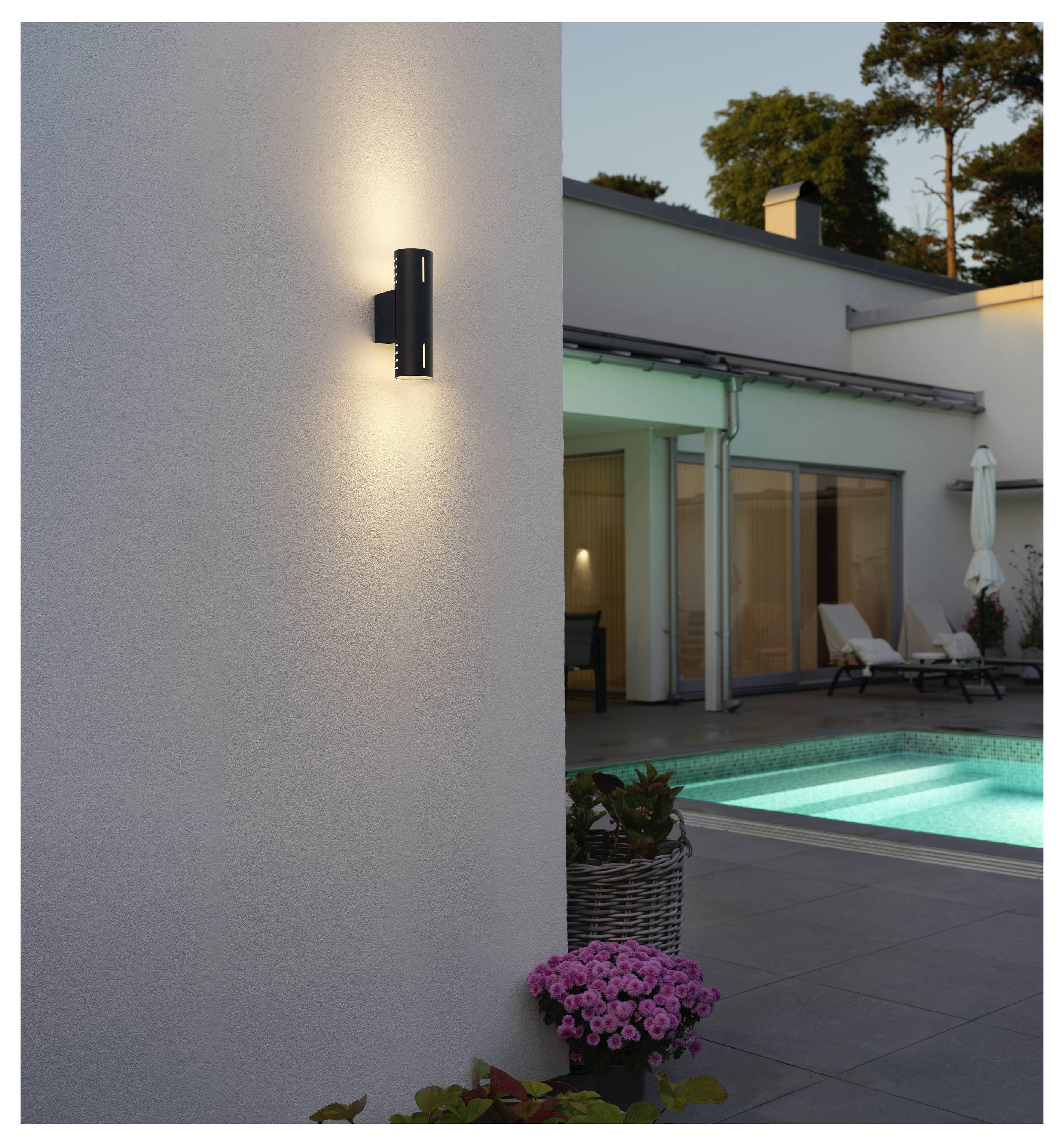 Nowoczesna lampa ścienna do użytku zewnętrznego oświetla strefę patio obok basenu, z widocznym w tle współczesnym domem podczas zmierzchu.