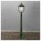 Zielona lampa w stylu vintage stoi na żwirowej ścieżce przy fakturowej szarej ścianie, oświetlając otoczenie.