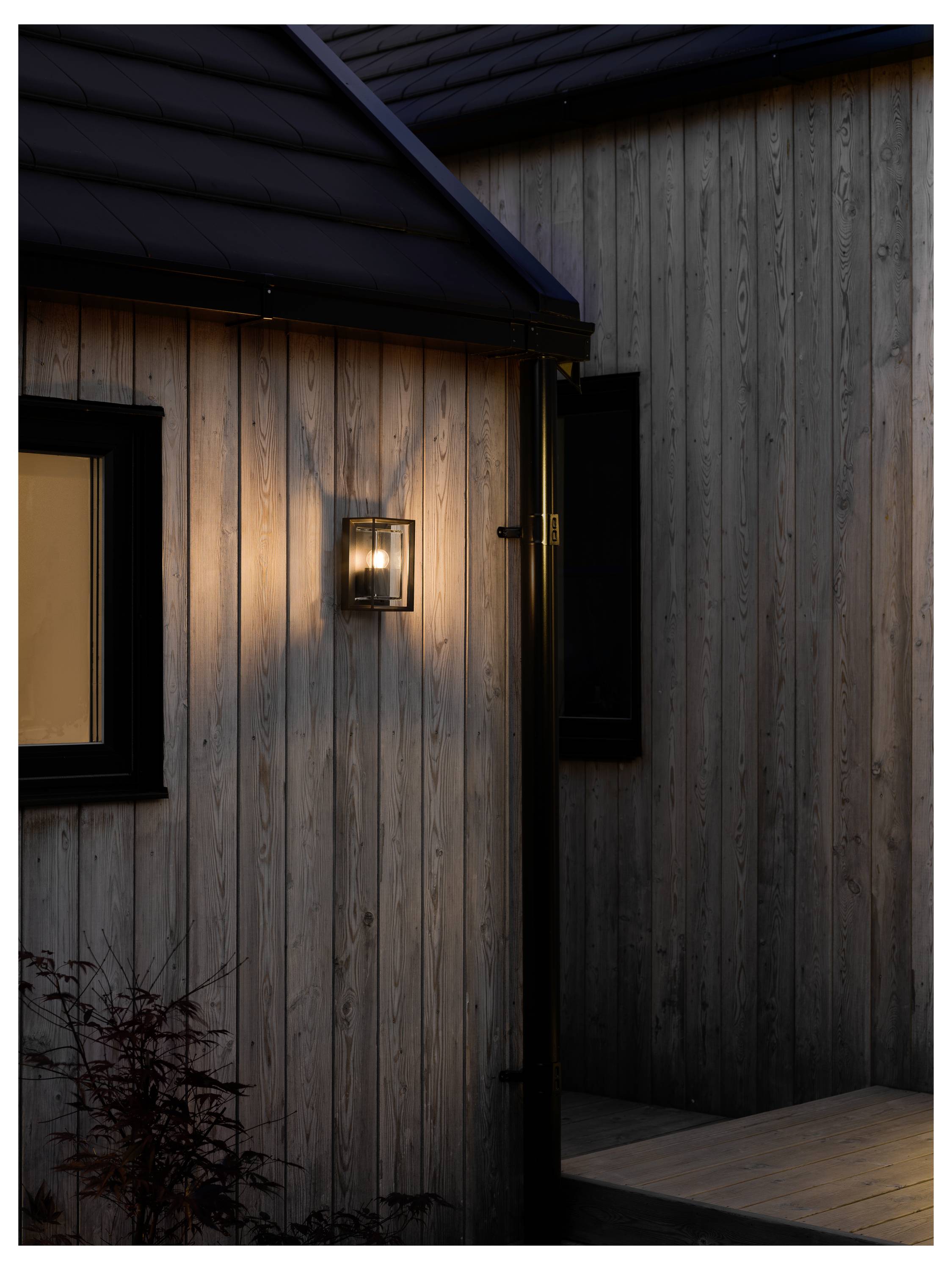 Drewniany exterior domu o zmierzchu z mocno świecącą lampą zamontowaną na ścianie, oświetlającą deskowanie przy oknie, tworząc ciepłą, przytulną atmosferę.