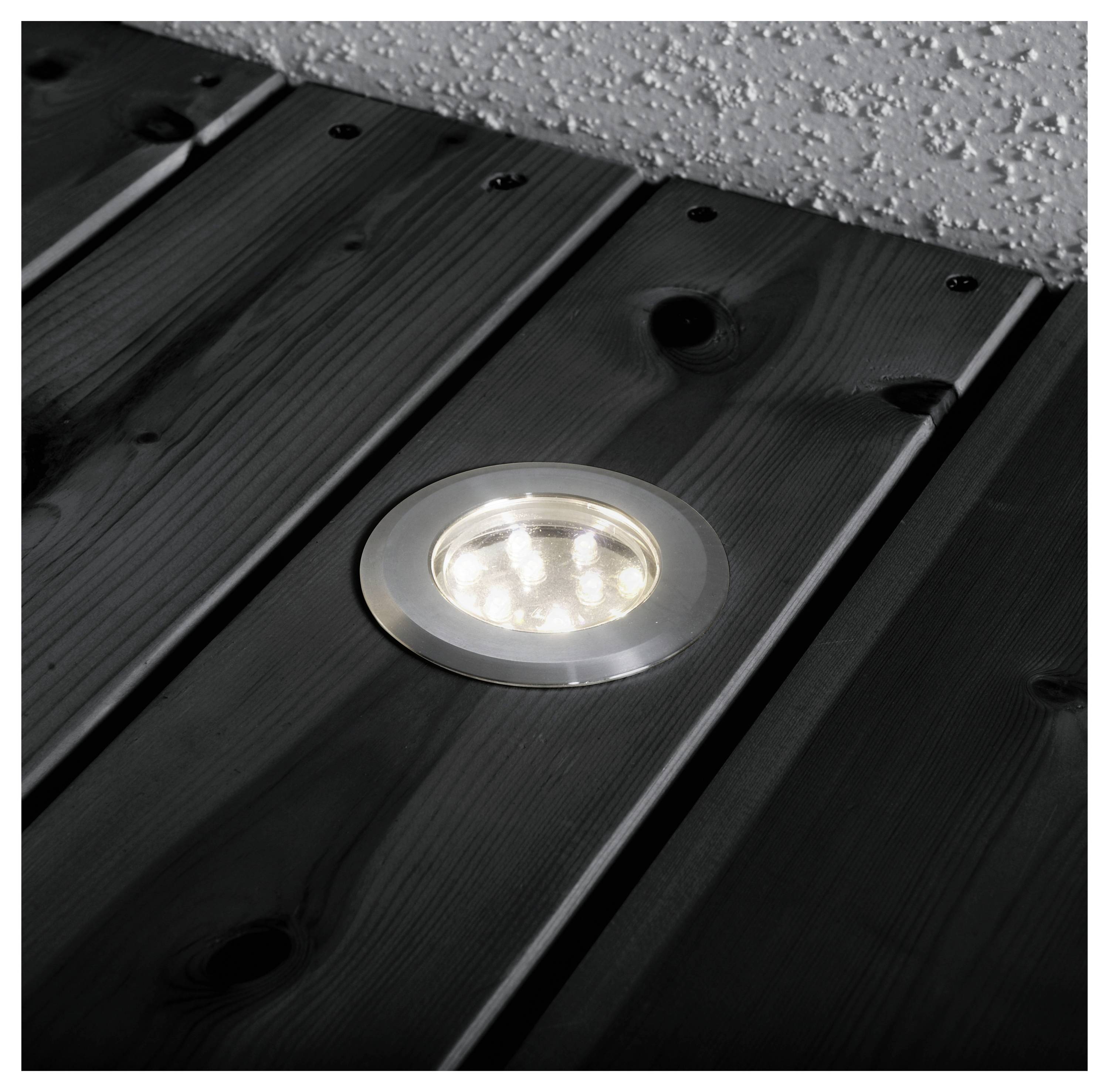 Wpuszczane okrągłe światło LED jest osadzone w ciemnych drewnianych deskach tarasowych, rzucając miękkie światło na deski i teksturowaną ścianę w pobliżu.