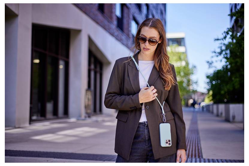 Osoba spaceruje na ulicy miasta w okularach przeciwsłonecznych i marynarce, trzymając smartfon na pasku przewieszonym przez szyję.