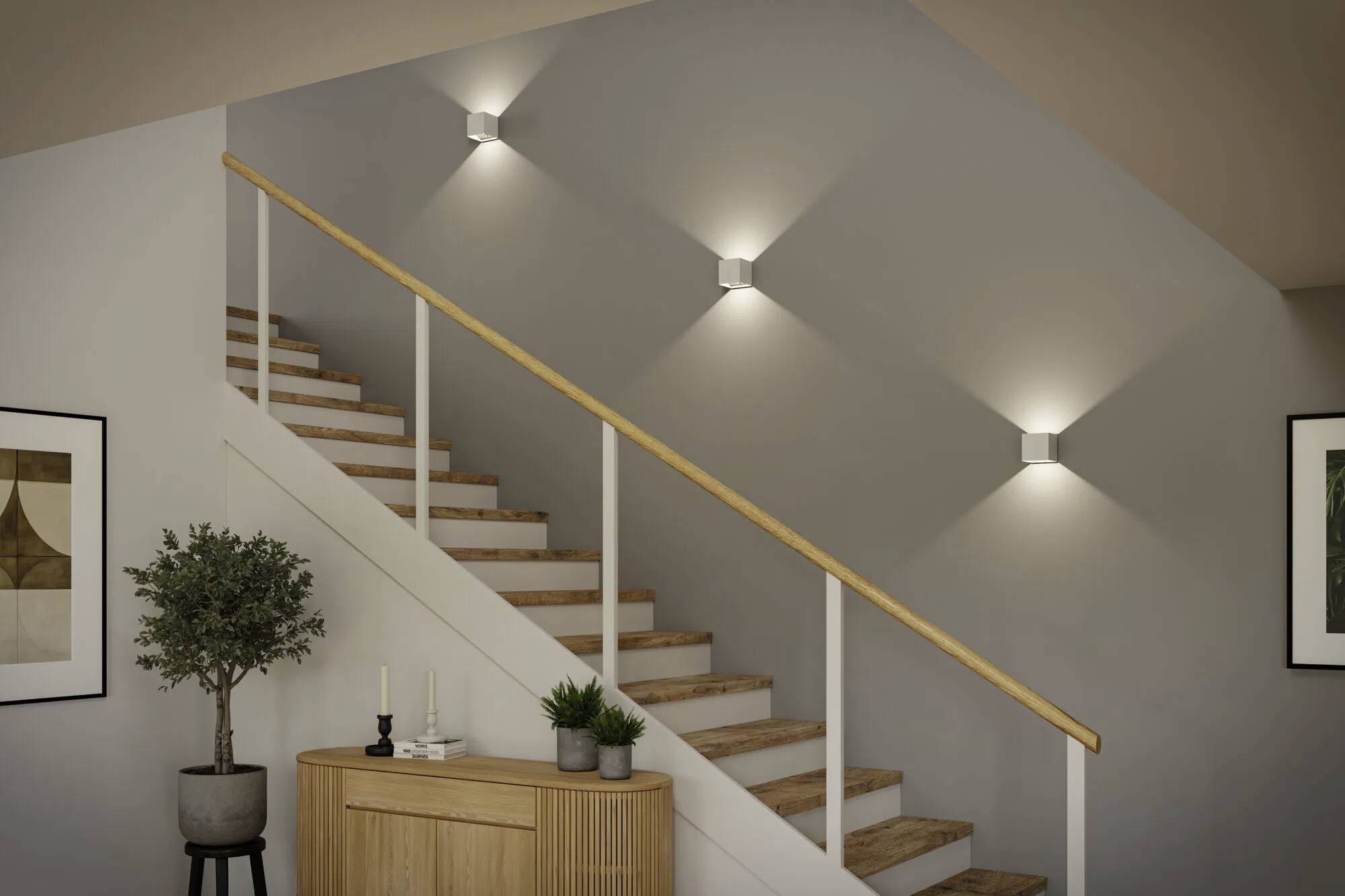 Drewniane schody z poręczą są oświetlone przez trzy lampy ścienne. U podstawy schodów stoi mała szafka z roślinami i świecami.
