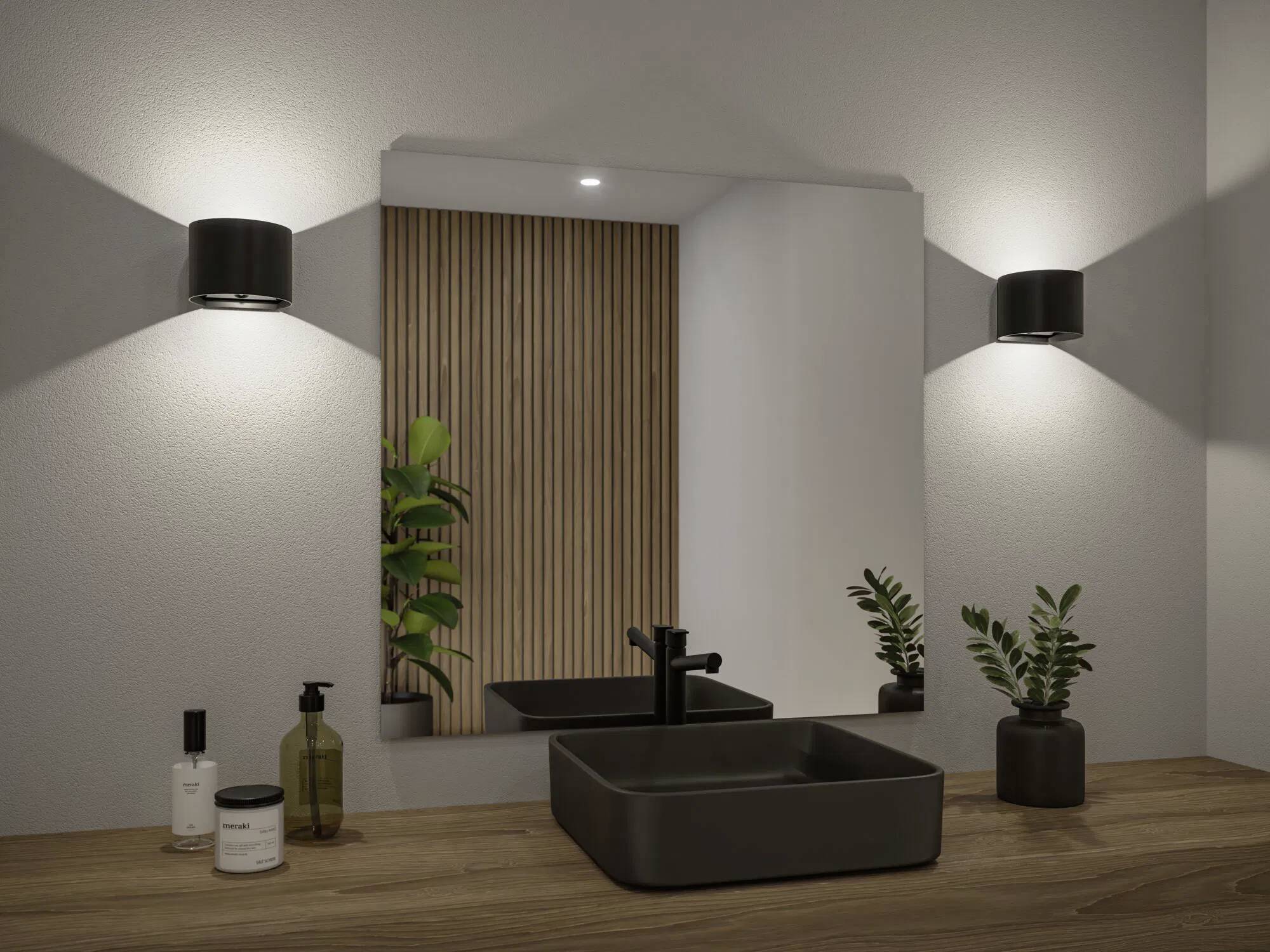 Nowoczesna szafka łazienkowa z prostokątnym lustrem, drewnianym blatem, czarną umywalką, baterią i doniczkowymi roślinami. Kinkiety rzucają miękkie światło.