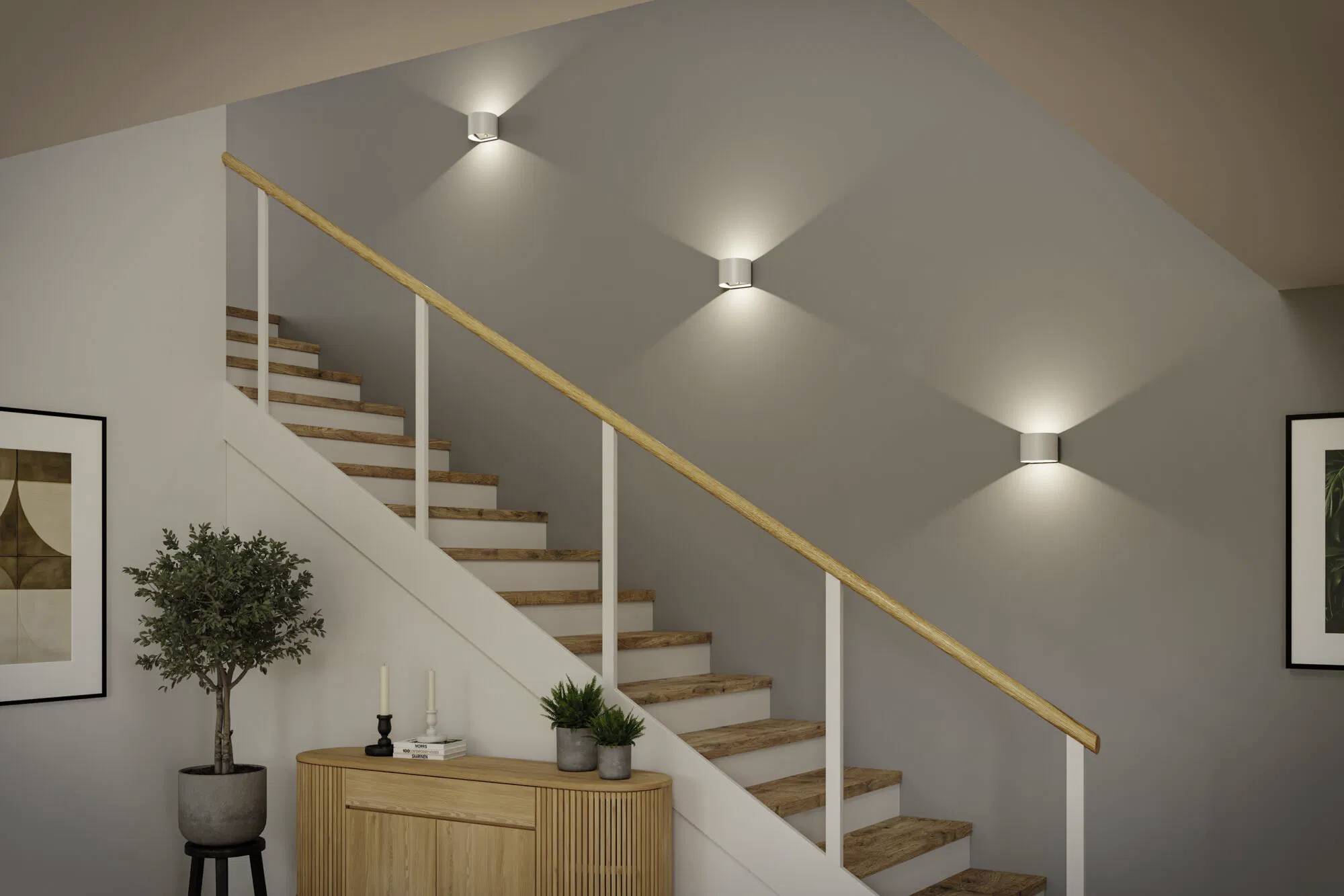 Nowoczesne drewniane schody z jasnobrązową poręczą i lampami zamocowanymi na ścianie. Mały stolik z roślinami i dekoracjami stoi poniżej.