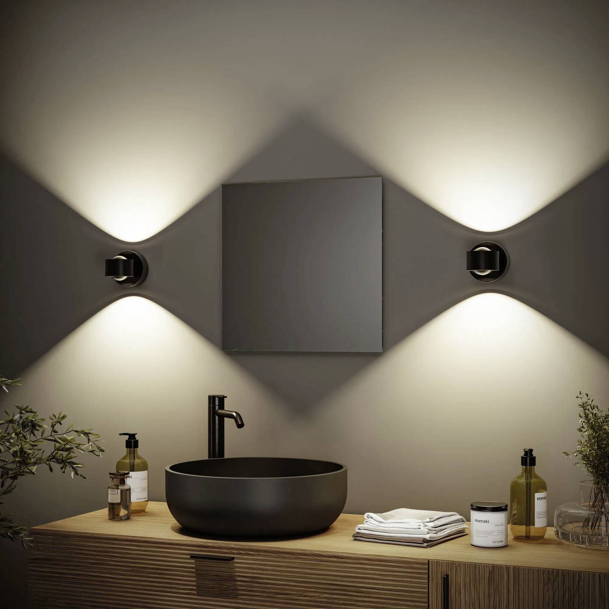 Nowoczesna łazienka z okrągłą czarną umywalką na drewnianym blacie, oświetlona kinkietami i ozdobiona roślinami oraz artykułami kosmetycznymi.
