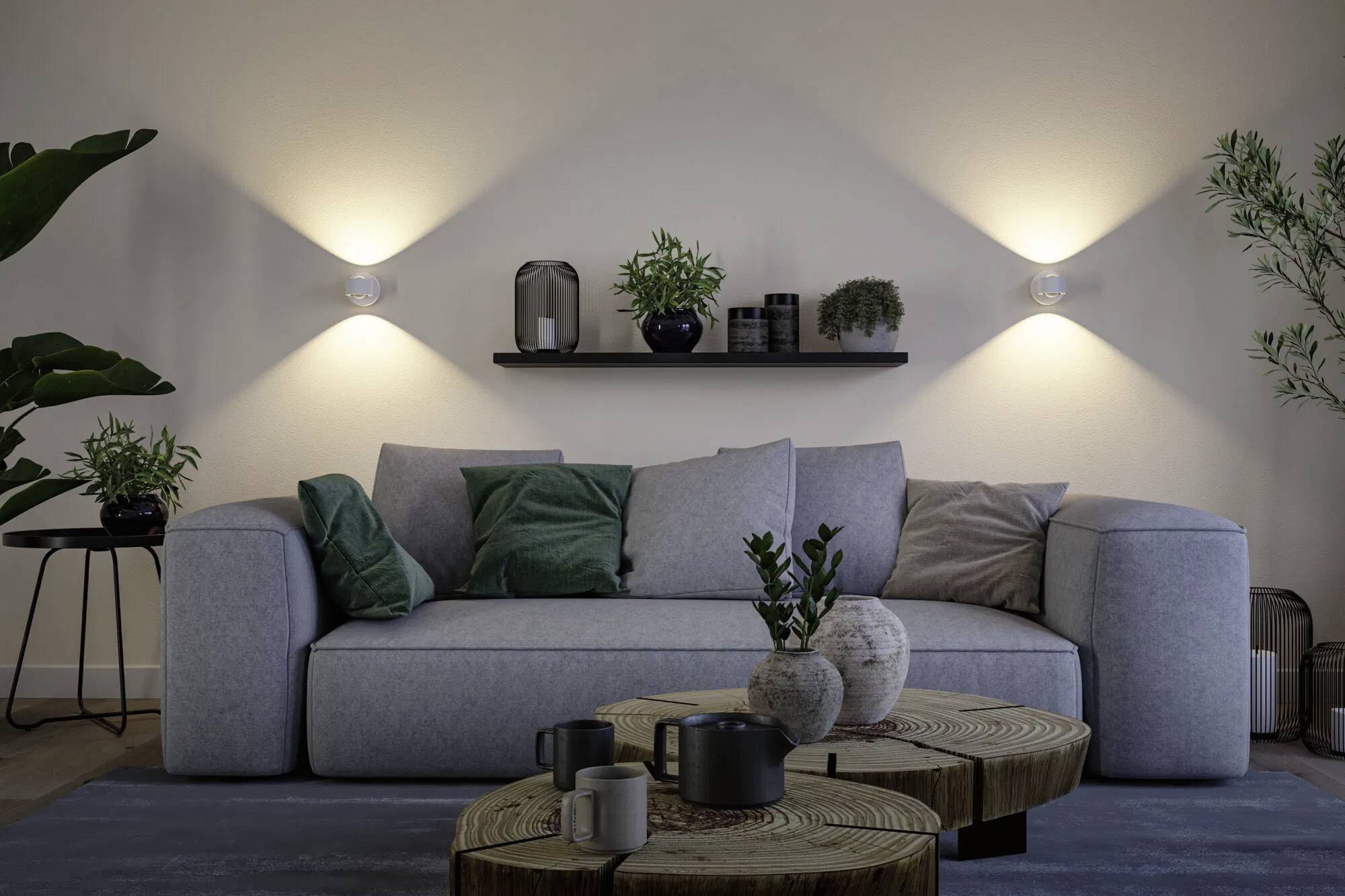 Przytulny salon z szarą sofą, zielonymi i szarymi poduszkami, drewnianymi stolikami kawowymi, roślinami i dekoracyjnymi kinkietami tworzącymi ciepłą atmosferę.