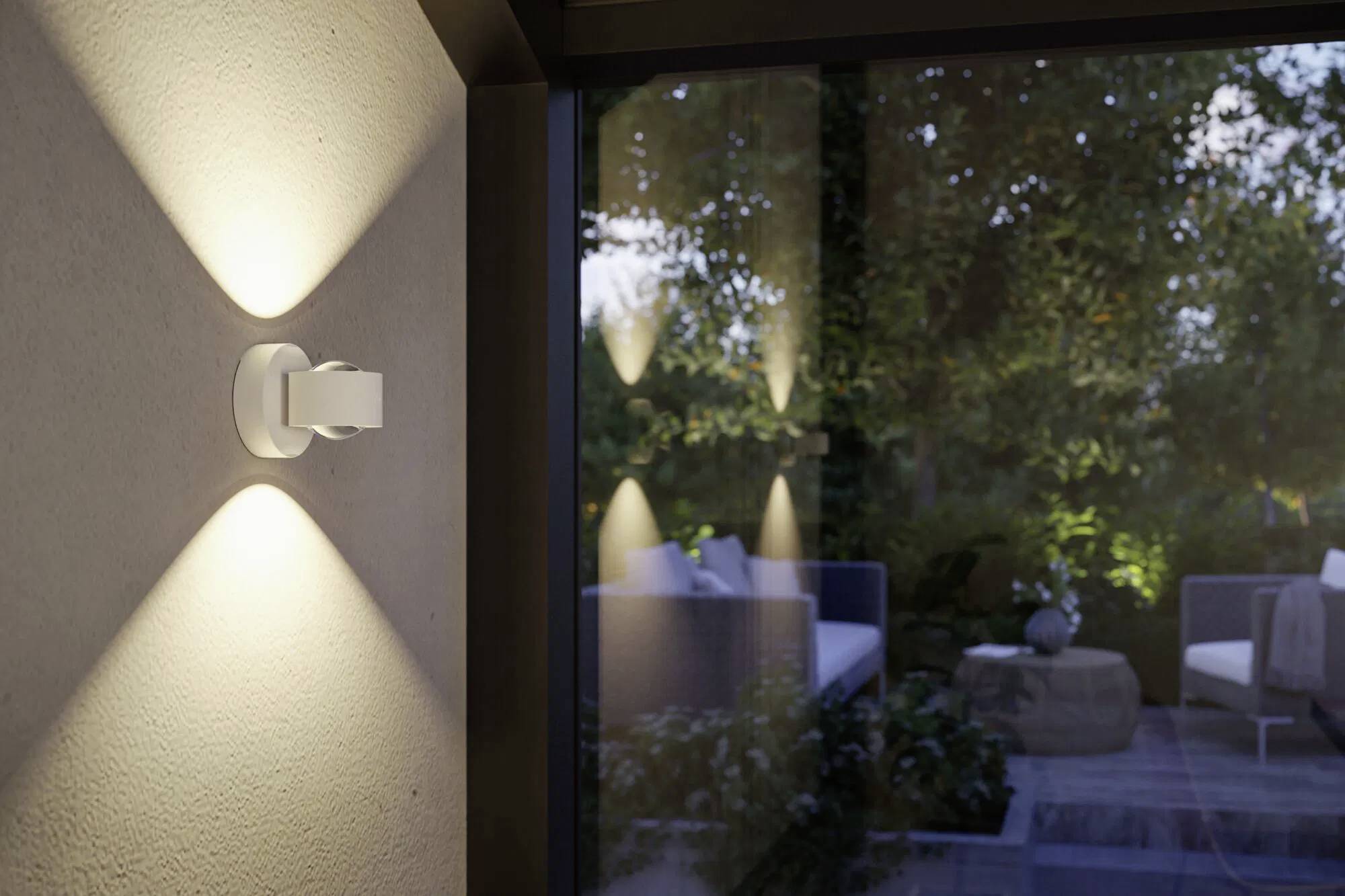 Nowoczesna lampa ścienna świeci na zewnętrznym tarasie, oświetlając siedziska i zieleń.