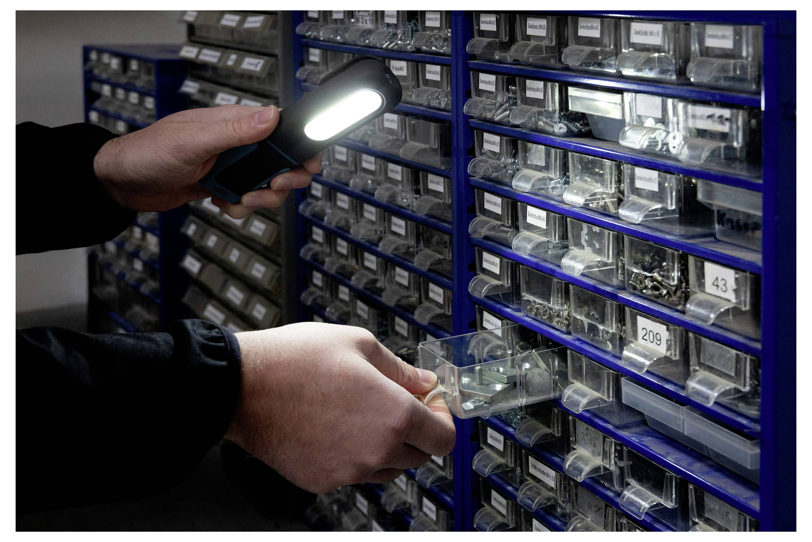 Osoba używa latarki do przeszukiwania opisanych szuflad wypełnionych małymi elementami sprzętowymi, podkreślając zorganizowany system przechowywania.