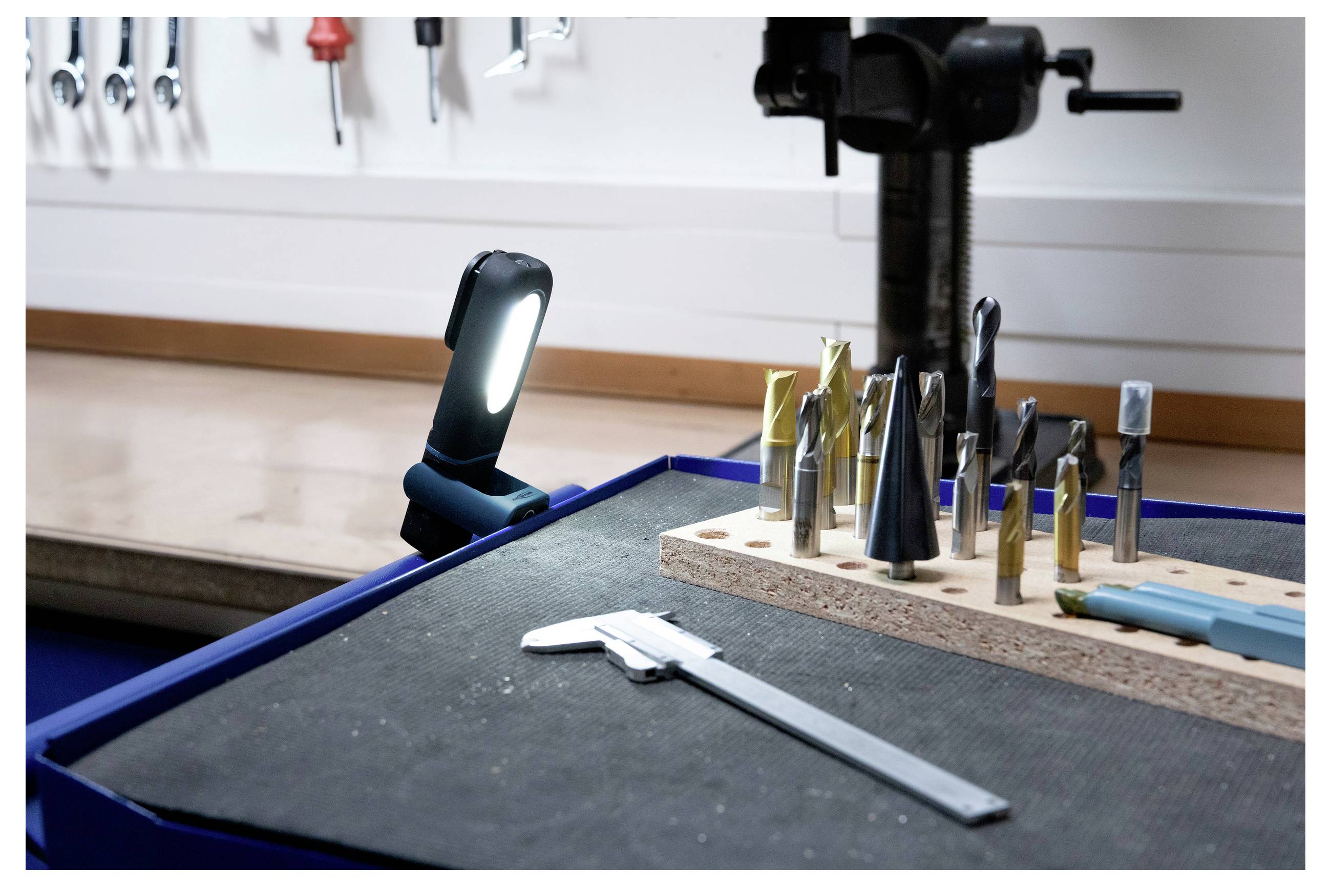 Warsztat z różnymi wiertłami ułożonymi na stojaku, jasna lampa oświetlająca przestrzeń oraz suwmiarka spoczywająca obok narzędzi.