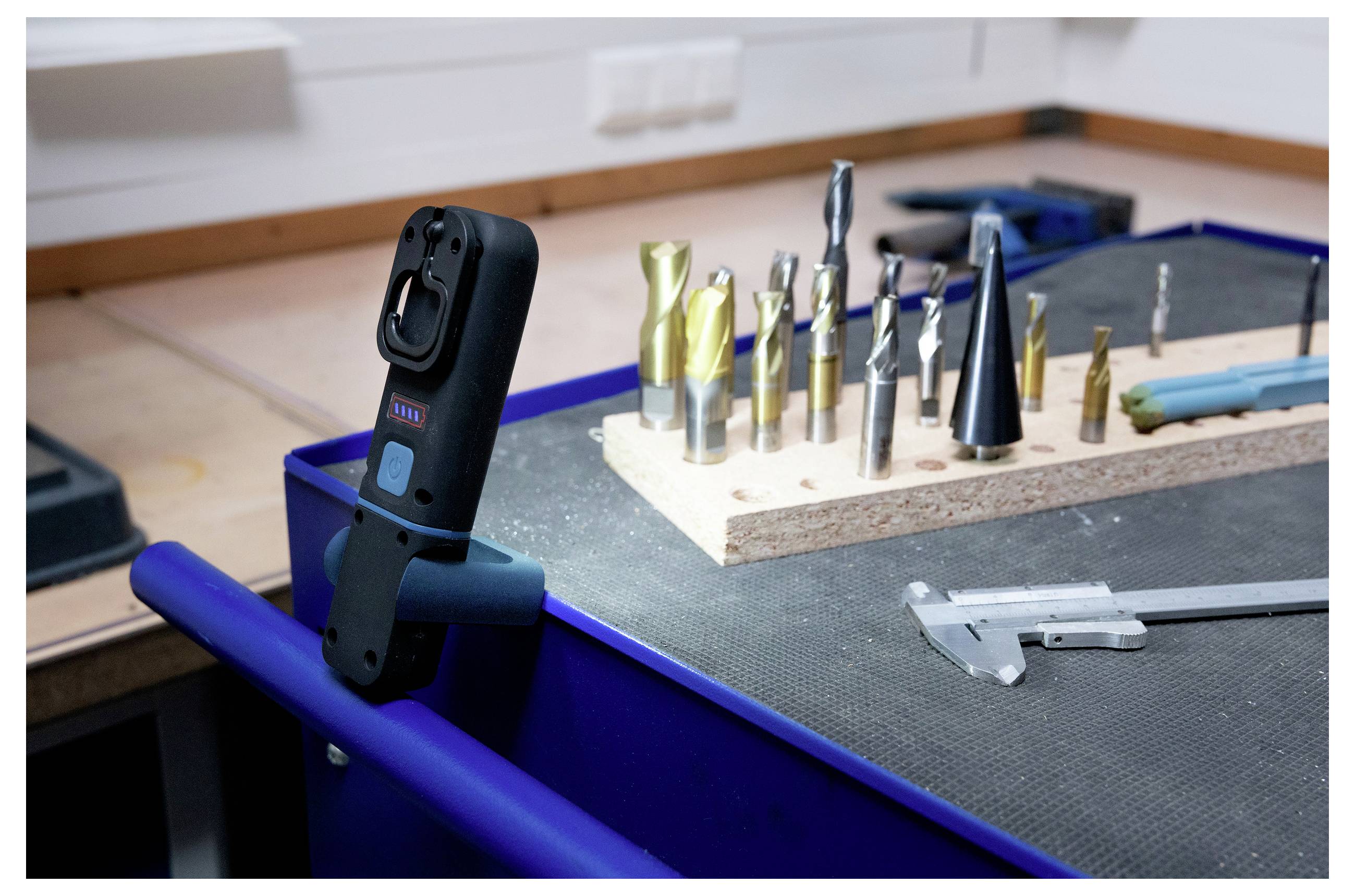 Stół warsztatowy prezentujący różne wiertła ułożone w drewnianym stojaku obok suwmiarki. Cyfrowy klucz dynamometryczny zamocowany w pobliżu.