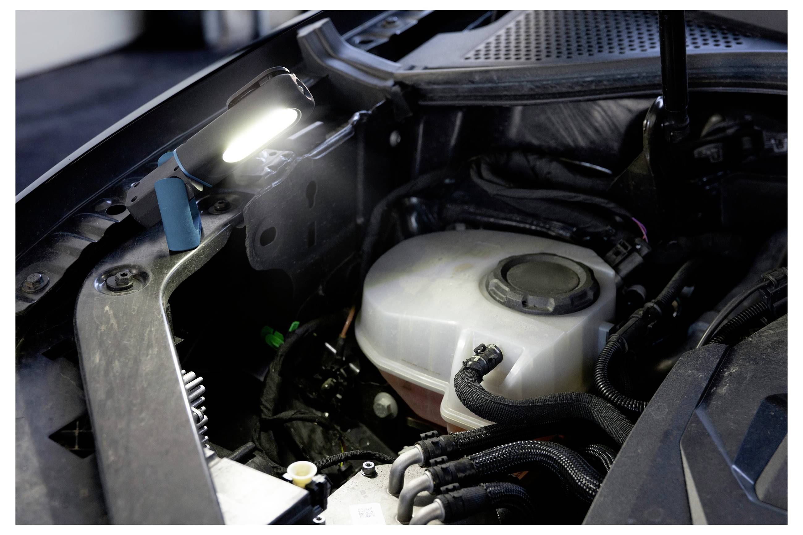 Podświetlona komora silnika samochodu prezentuje zbiornik płynu chłodzącego oraz różne przewody, z lampą oświetlającą obszar do przeprowadzenia czynności serwisowych.