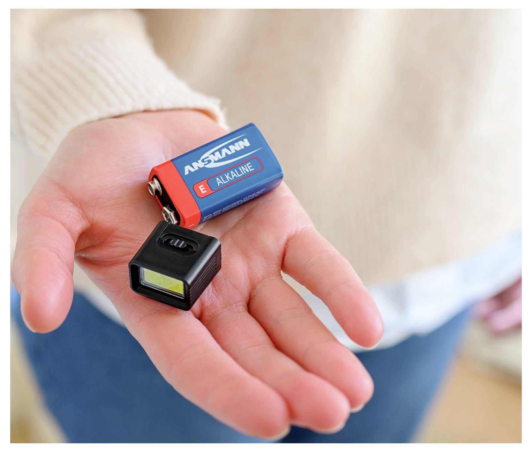 Ręka trzymająca baterię dziewięciovoltową i małe urządzenie elektroniczne, prawdopodobnie tester, z rozmytym tłem.