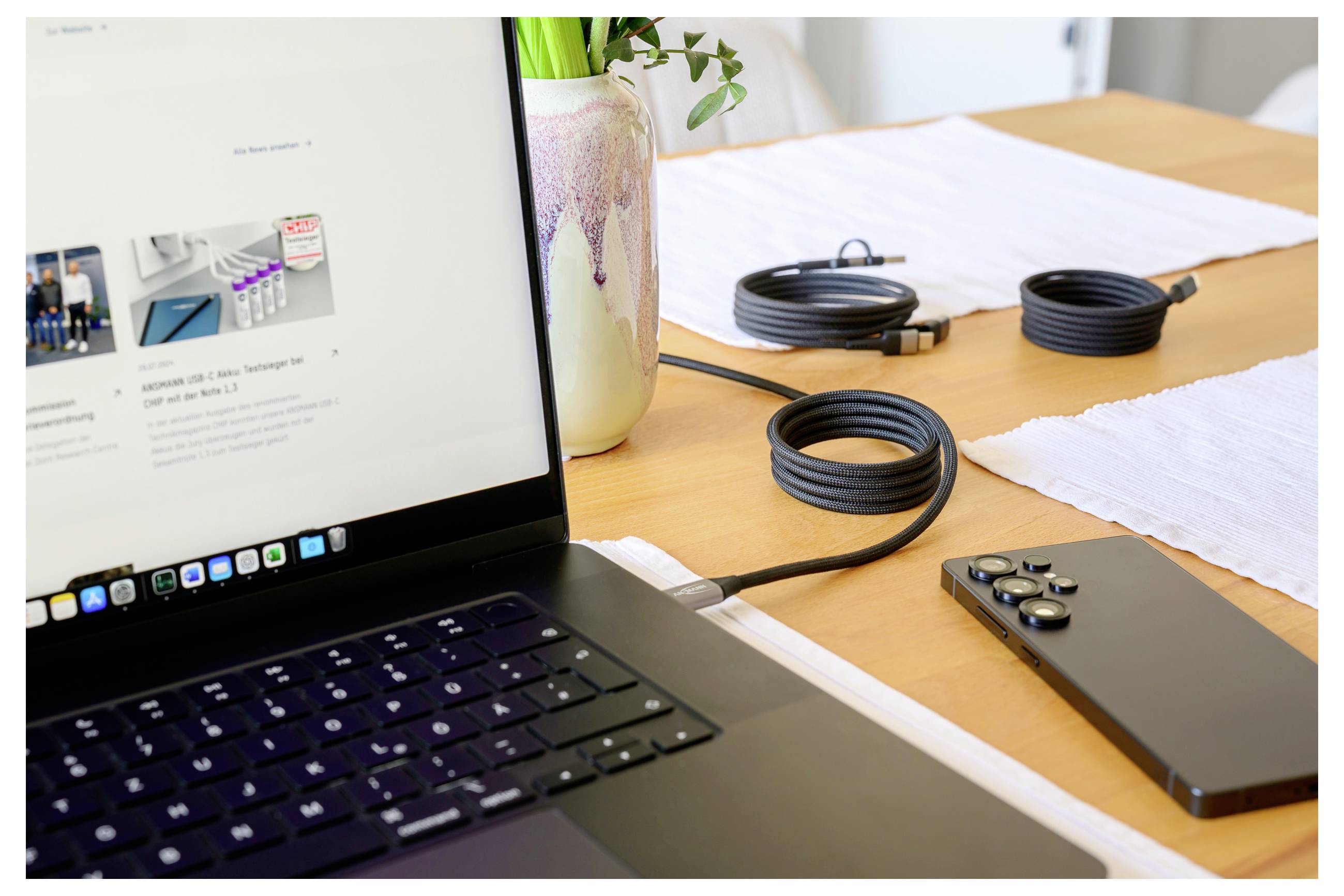 Laptop na stole podłączony kablem do smartfona, z kręconymi przewodami i wazonem z zielonymi liśćmi w tle.