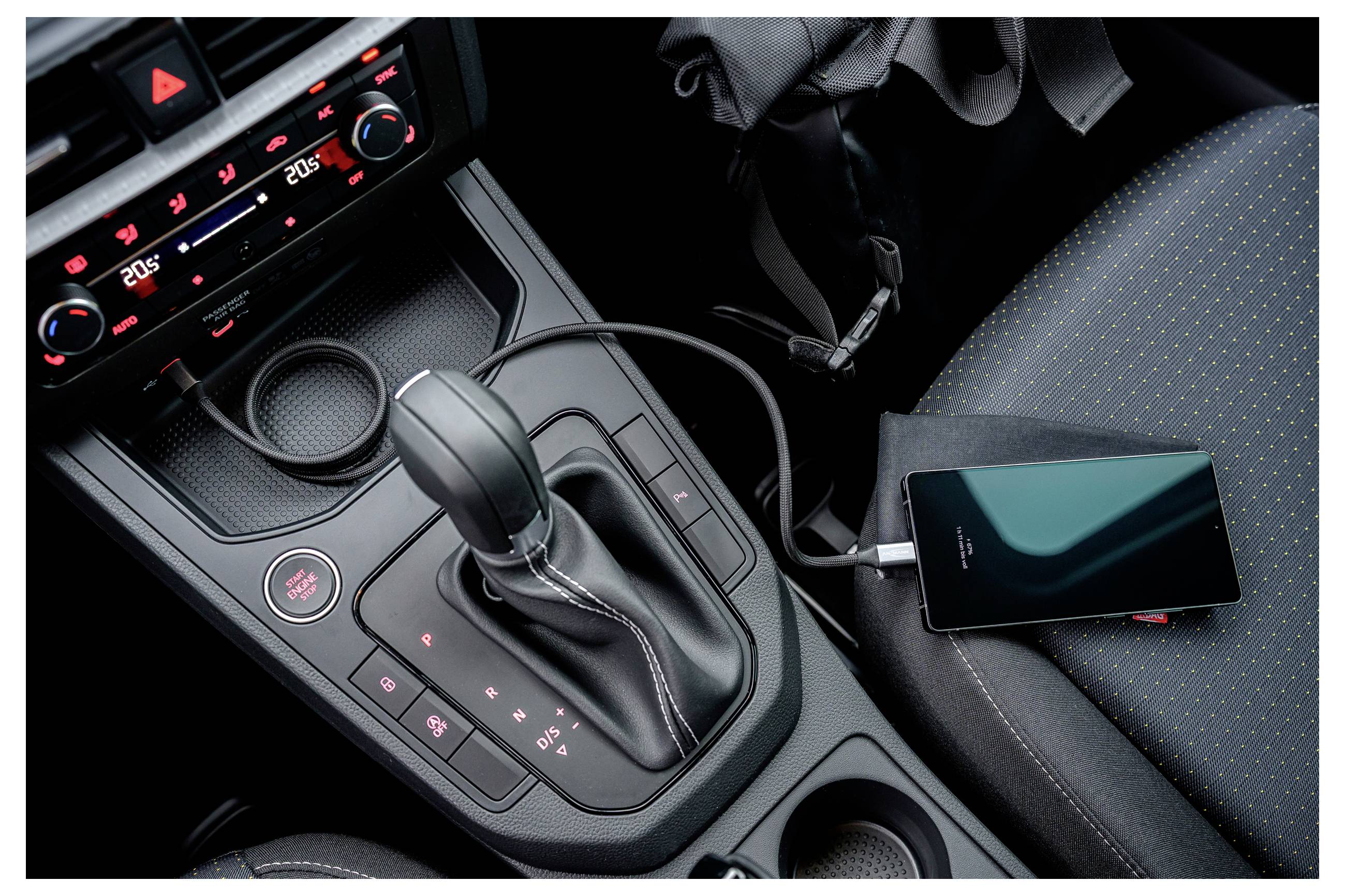 Smartfon ładujący się w samochodzie, podłączony kablem. Widoczna dźwignia zmiany biegów i elementy sterowania na desce rozdzielczej, z wyświetlaczem temperatury.