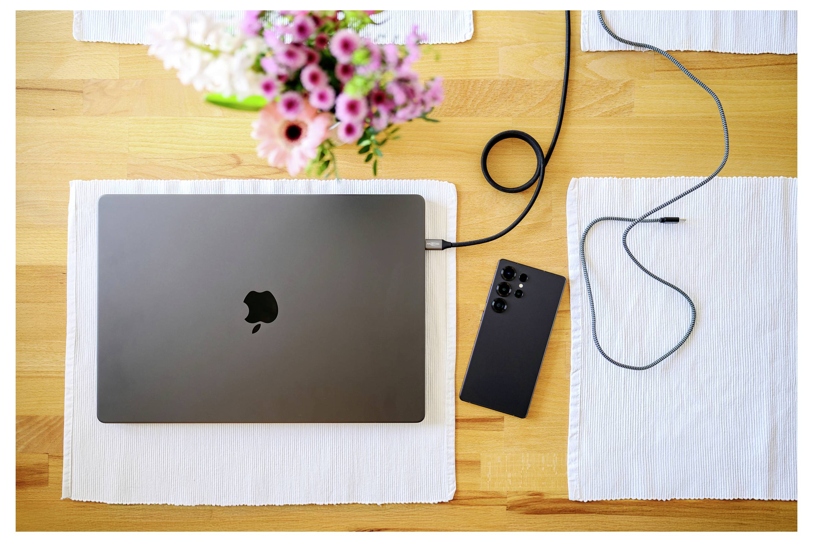 Laptop ładuje się na drewnianym stole, otoczony smartfonem, kablem ładującym i wazonem z różowymi kwiatami.