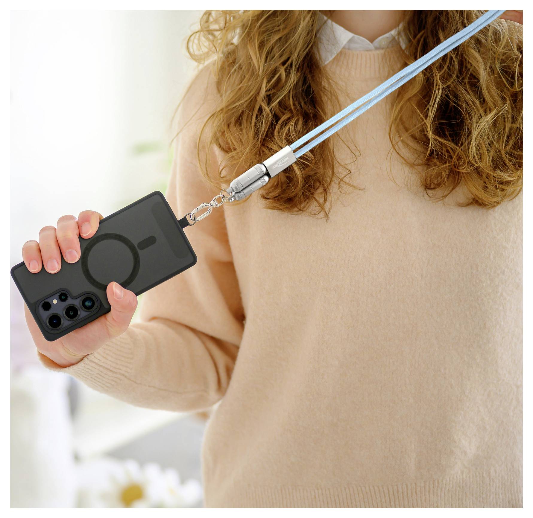 Osoba ubrana w beżowy sweter trzyma czarnego smartfona podłączonego do niebieskiego smyczy, prezentując opcję noszenia bez użycia rąk.