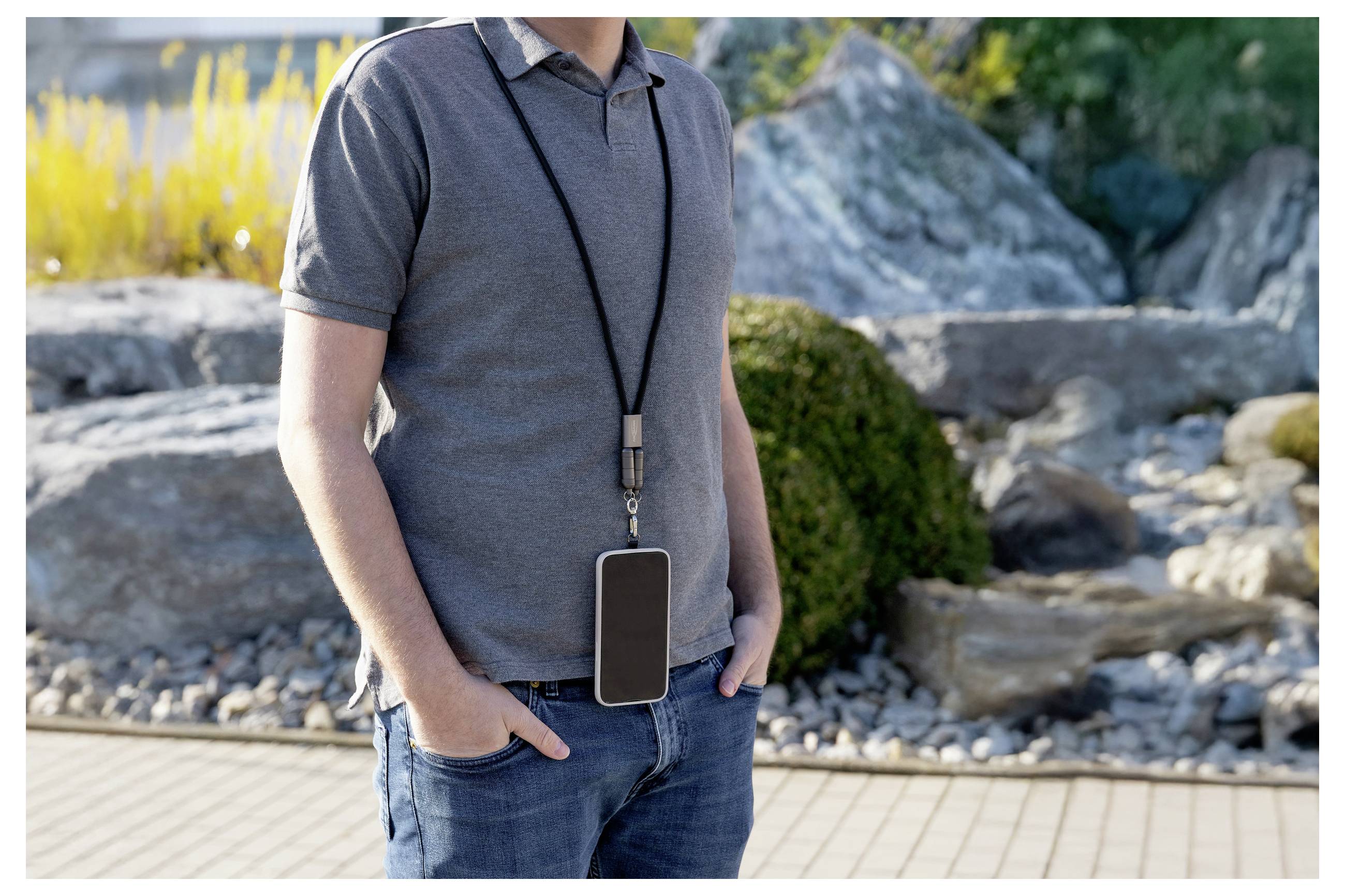 Mężczyzna w szarej koszulce polo i dżinsach stoi na dworze z rękami w kieszeniach, noszący duży smartfon na smyczy zawieszonej na szyi.