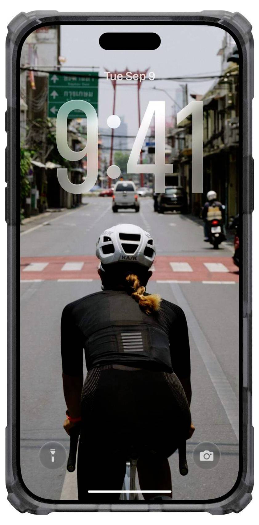 Rowerzysta w kasku jedzie w kierunku czerwonego przejścia dla pieszych, a w tle widać jasne niebo i budynki uliczne, oglądane przez ekran smartfona wskazujący godzinę 9:41.