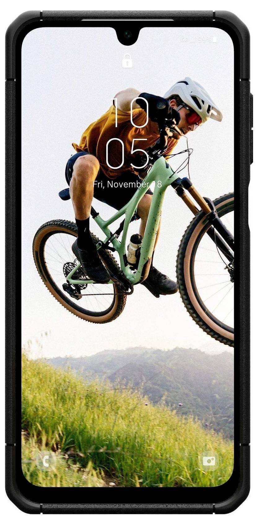 Rowerzysta górski w powietrzu, wykonujący skok na szlaku, z błękitnym niebem i oddalonymi wzgórzami w tle.