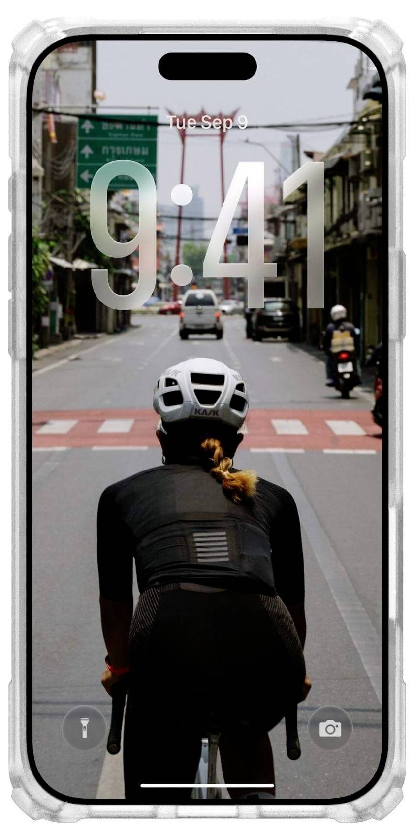 Osoba w kasku jedzie na rowerze pustą ulicą miejską, widok od tyłu. Przed nią widoczne są sygnalizatory świetlne i budynki.