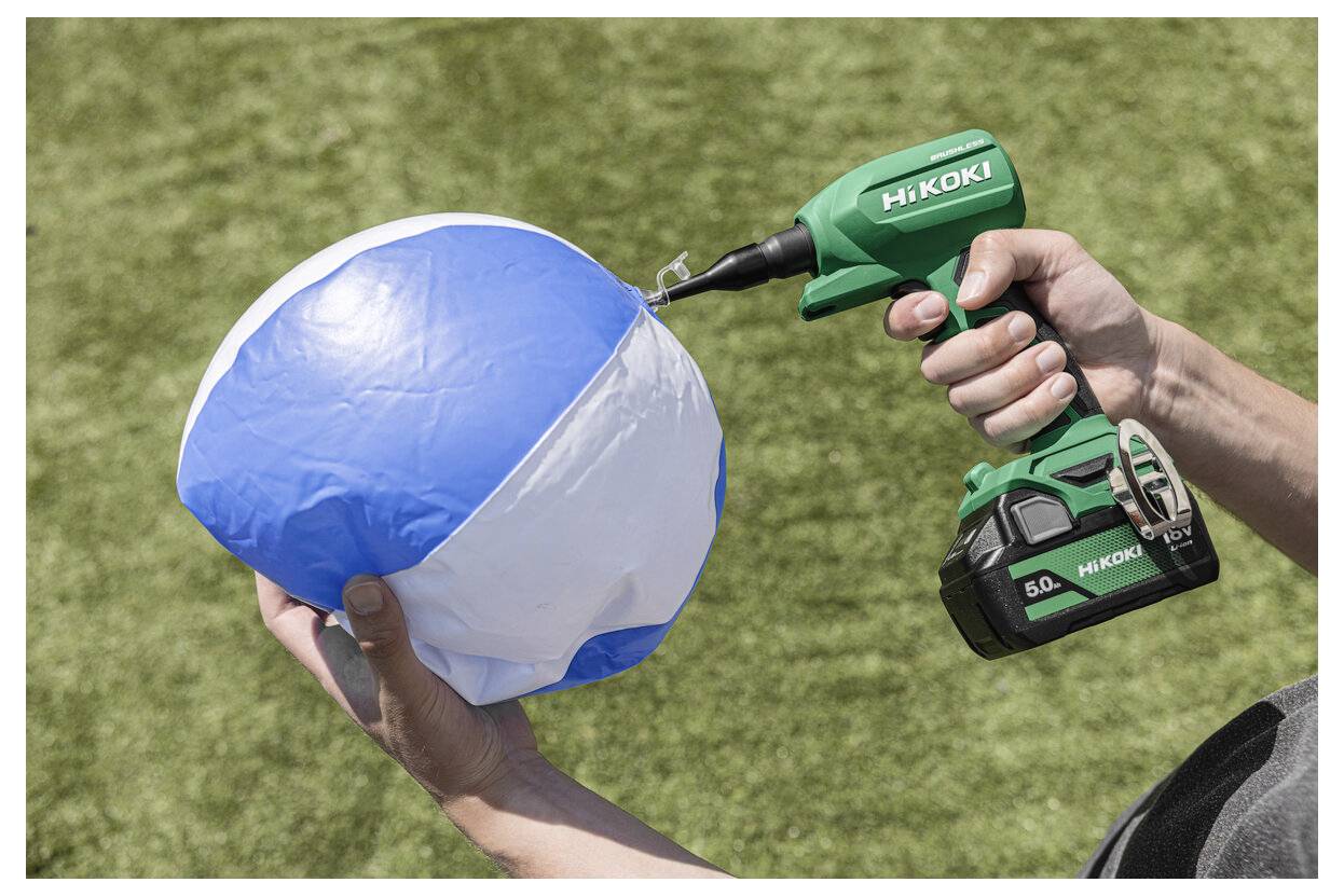 Osoba używa zielonej wiertarki akumulatorowej na zdmuchanej niebiesko-białej piłce plażowej, sugerując humorystyczne lub niekonwencjonalne działanie na trawie.