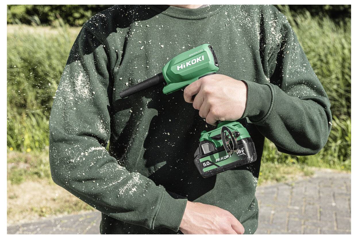 Osoba trzymająca zielone elektronarzędzie HiKOKI z baterią, pokryte trocinami, stojąca na dworze w słoneczny dzień.