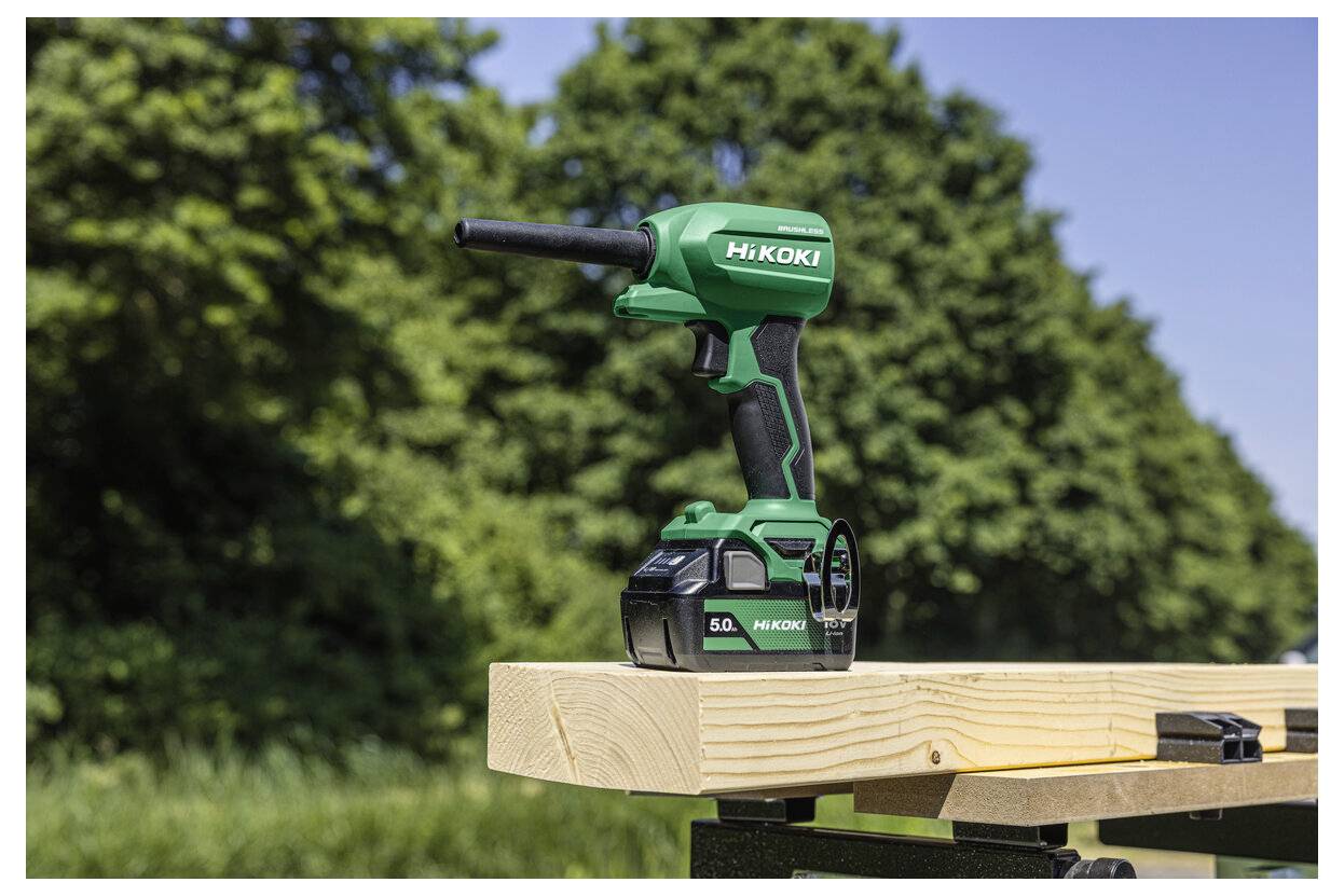 Zielone elektronarzędzie HiKOKI, prawdopodobnie akumulatorowy wkrętak udarowy, spoczywa na drewnianej desce na wolnym powietrzu, w tle widać drzewa.