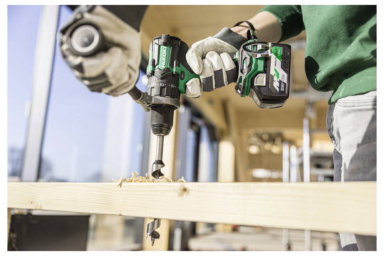 Osoba używa zielonej akumulatorowej wiertarki do wywiercenia otworu w drewnianej desce w jasnym warsztacie, nosząc rękawice ochronne dla bezpieczeństwa.