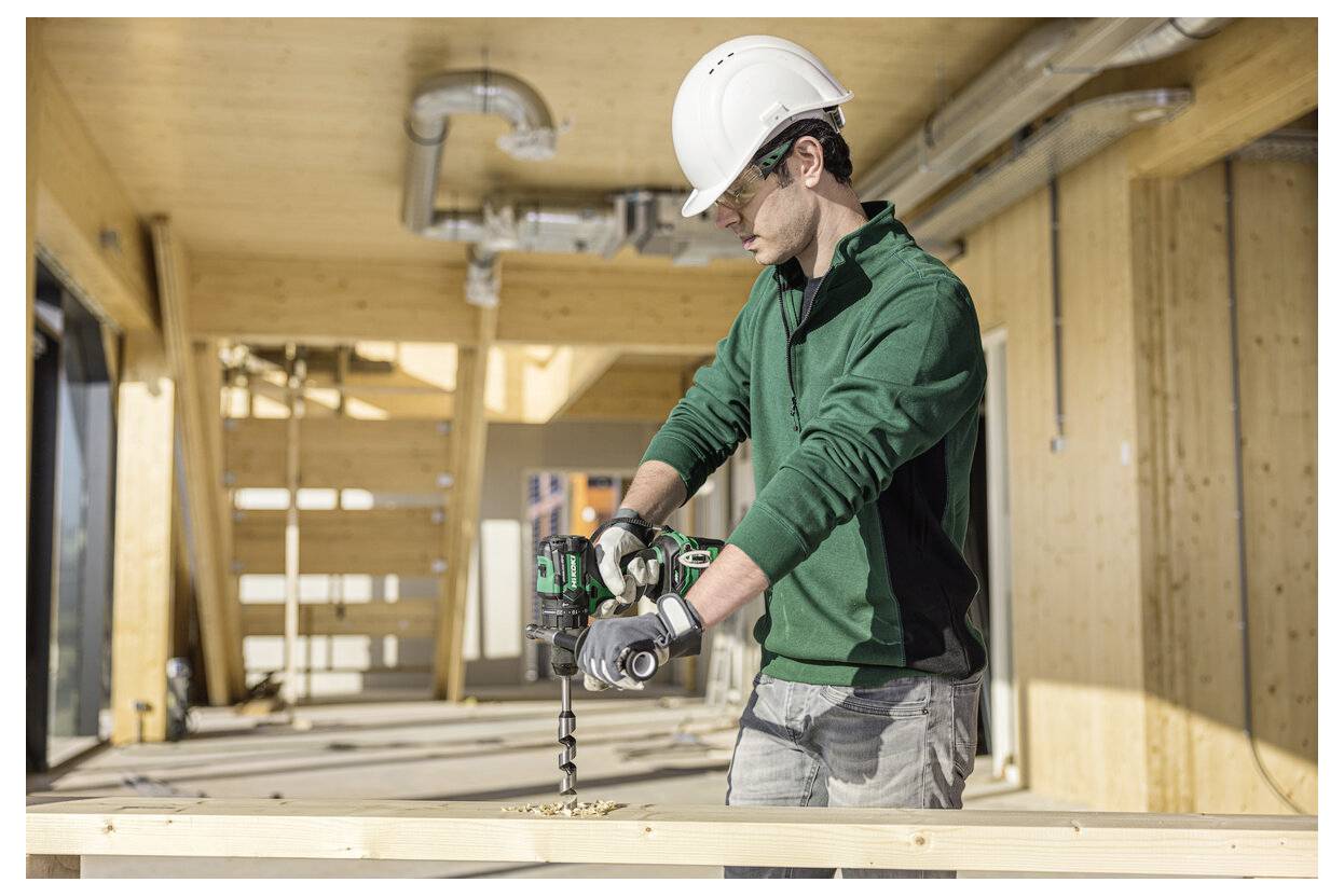 Robotnik budowlany w zielonej kurtce i kasku wwierca się w drewnianą belkę na placu budowy.