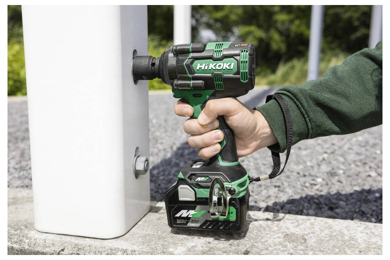 Osoba używa bezprzewodowego klucza udarowego na metalowej śrubie przymocowanej do białego słupa na wolnym powietrzu, z żwirową powierzchnią i zielenią w tle.