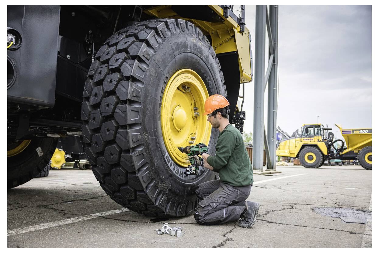 Osoba w pomarańczowym kasku naprawia duży żółty opona na maszynie budowlanej. W tle widoczny jest podobny pojazd.