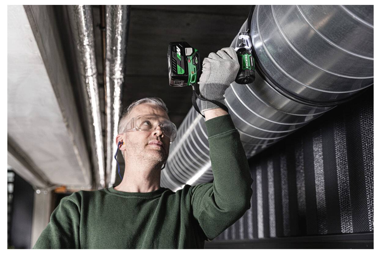 Osoba ubrana w okulary ochronne i rękawice używa wiertarki elektrycznej do instalacji lub naprawy metalowego przewodu wentylacyjnego w środowisku przemysłowym.