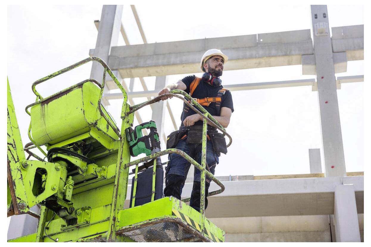 Robotnik budowlany w kasku ochronnym i pozostałym sprzęcie ochronnym stoi na zielonym podnośniku nożycowym na terenie budowy, obserwując postęp wznoszenia budynku.