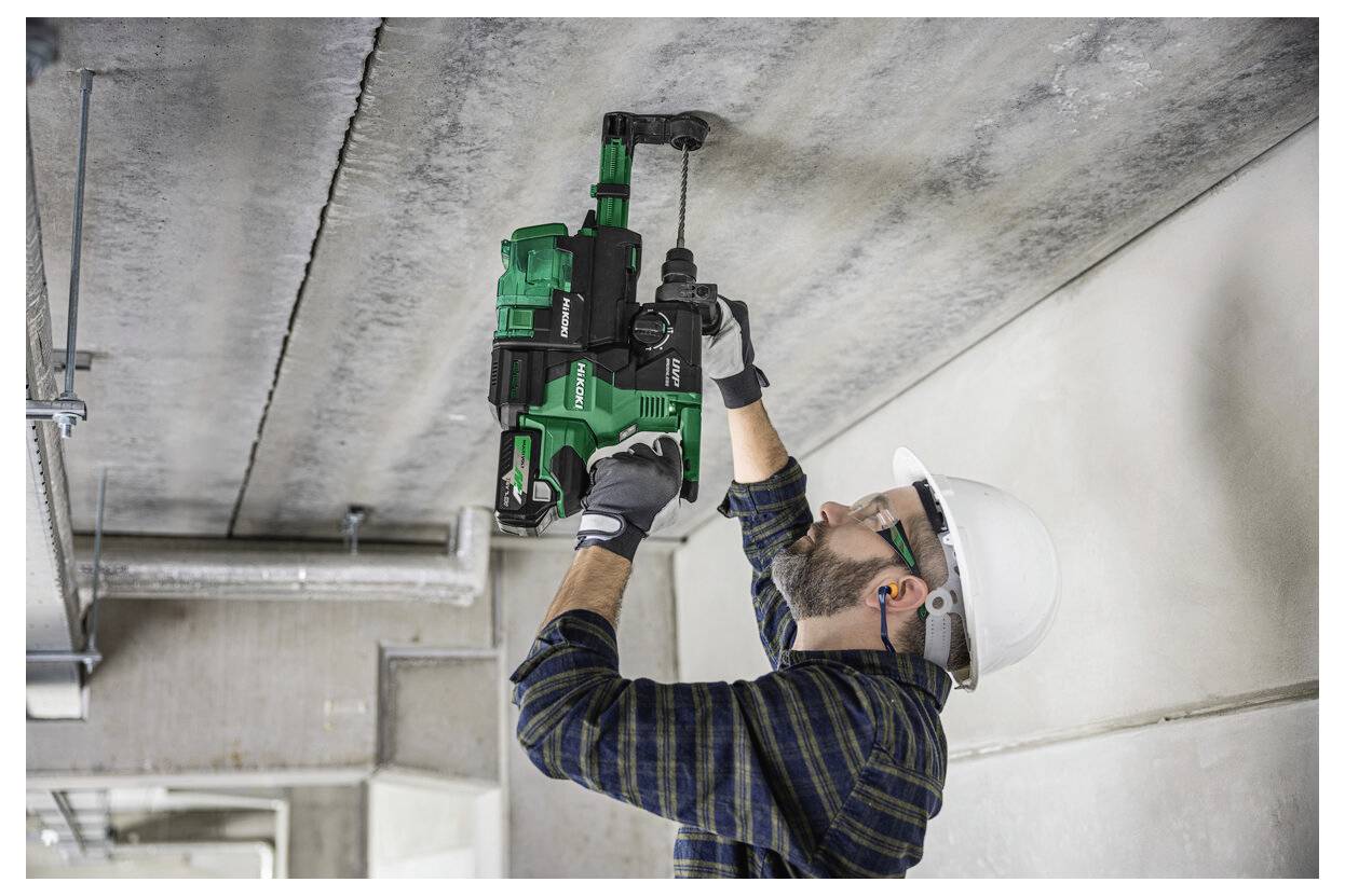 Robotnik budowlany w sprzęcie ochronnym używa zielonej wiertarko-młotowiertarki na betonowym suficie, koncentrując się na precyzji i stabilności.