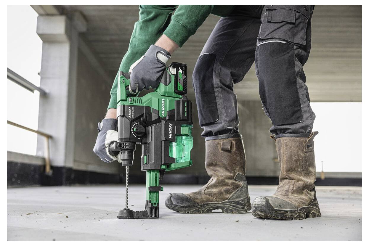 Osoba w rękawicach roboczych obsługuje zieloną wiertarkę udarową na betonowej podłodze, prezentując działalność budowlaną lub remontową.