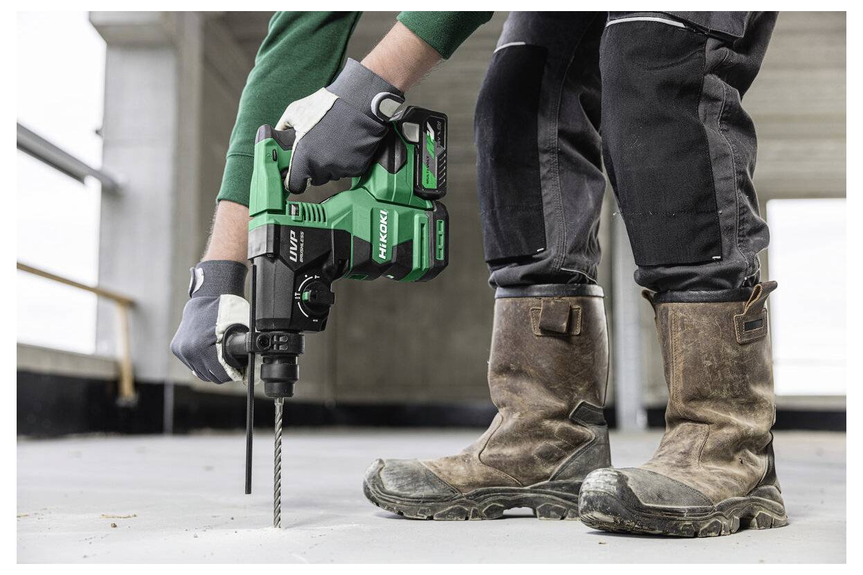 Osoba ubrana w rękawice i buty używa zielonej wiertarki udarowej na betonowej podłodze w niedokończonym budynku.