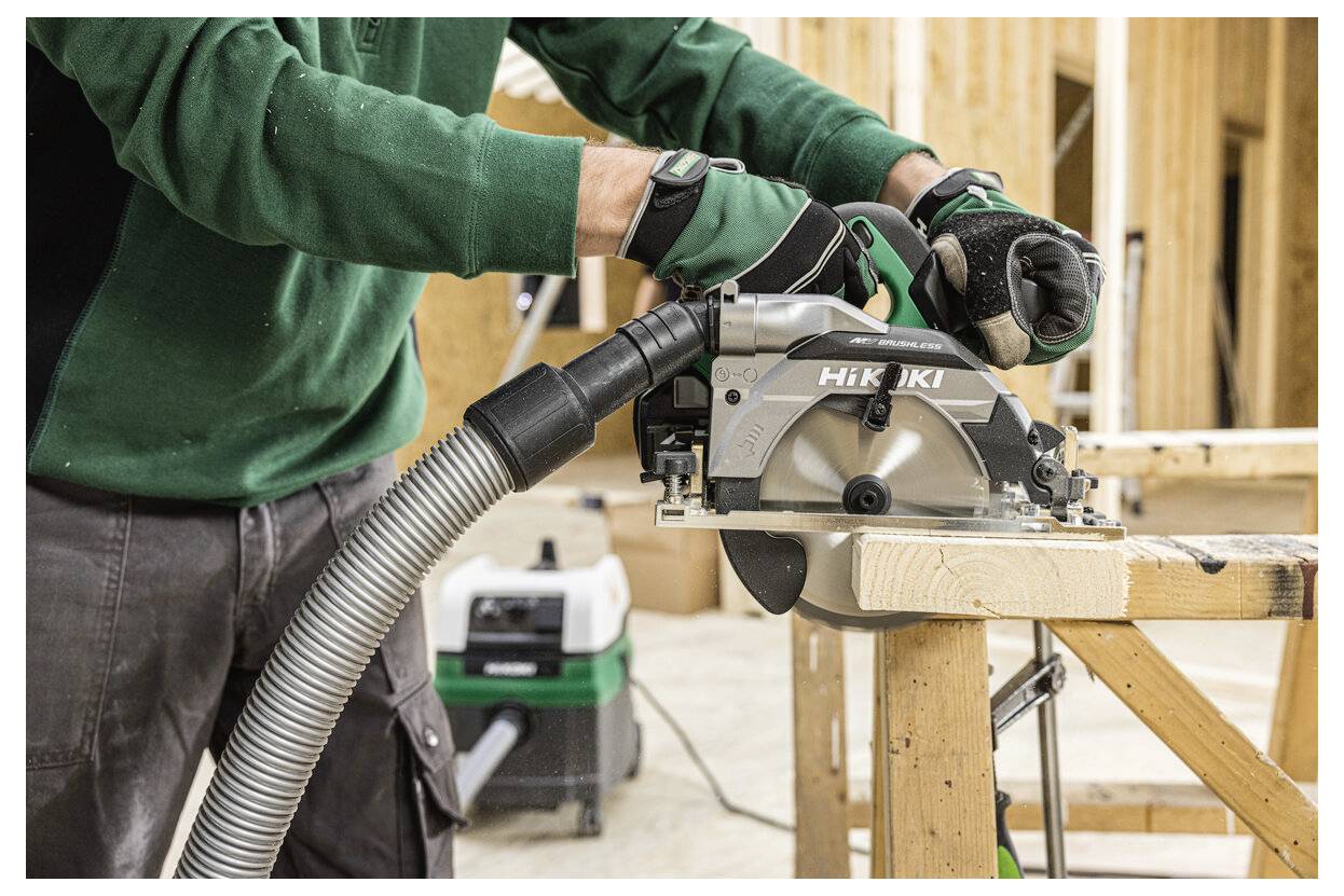 Osoba używająca piły tarczowej do cięcia drewna, ubrana w zielone rękawice i zieloną górę, w warsztacie z innymi narzędziami w tle.