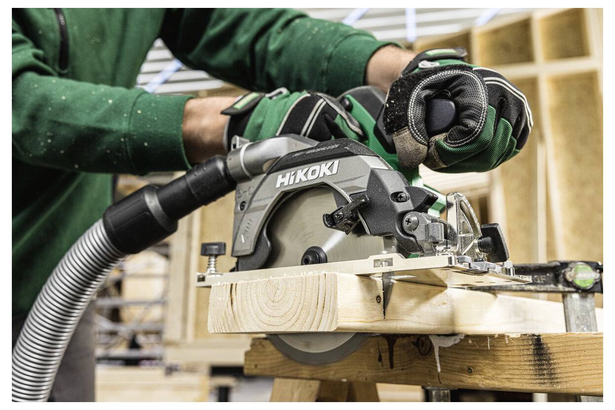 Osoba w zielonej kurtce używająca piły tarczowej do cięcia drewna, z troczynami zbieranymi przez podłączony wąż odkurzacza w warsztacie.