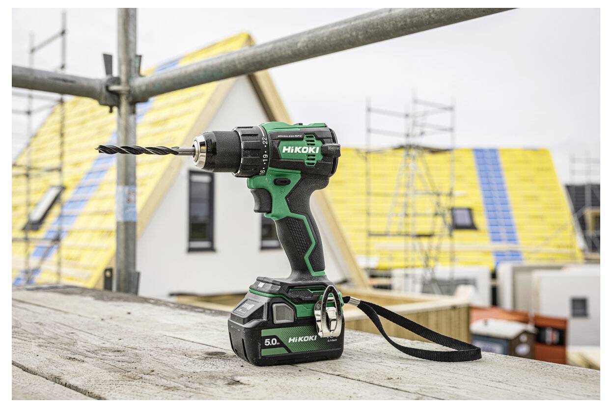 Akumulatorowa wiertarka z wiertłem leży na drewnianej desce na placu budowy, w tle częściowo wybudowane domy.