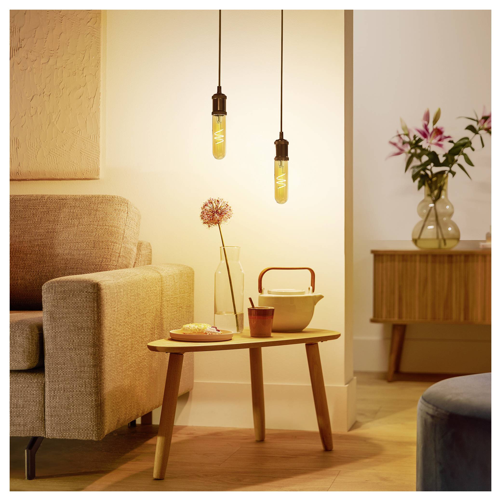 Przytulny kącik salonu z beżową sofą, wiszącymi żarówkami Edison, drewnianym stolikiem kawowym z różowym kwiatem w wazonie oraz kredensu.