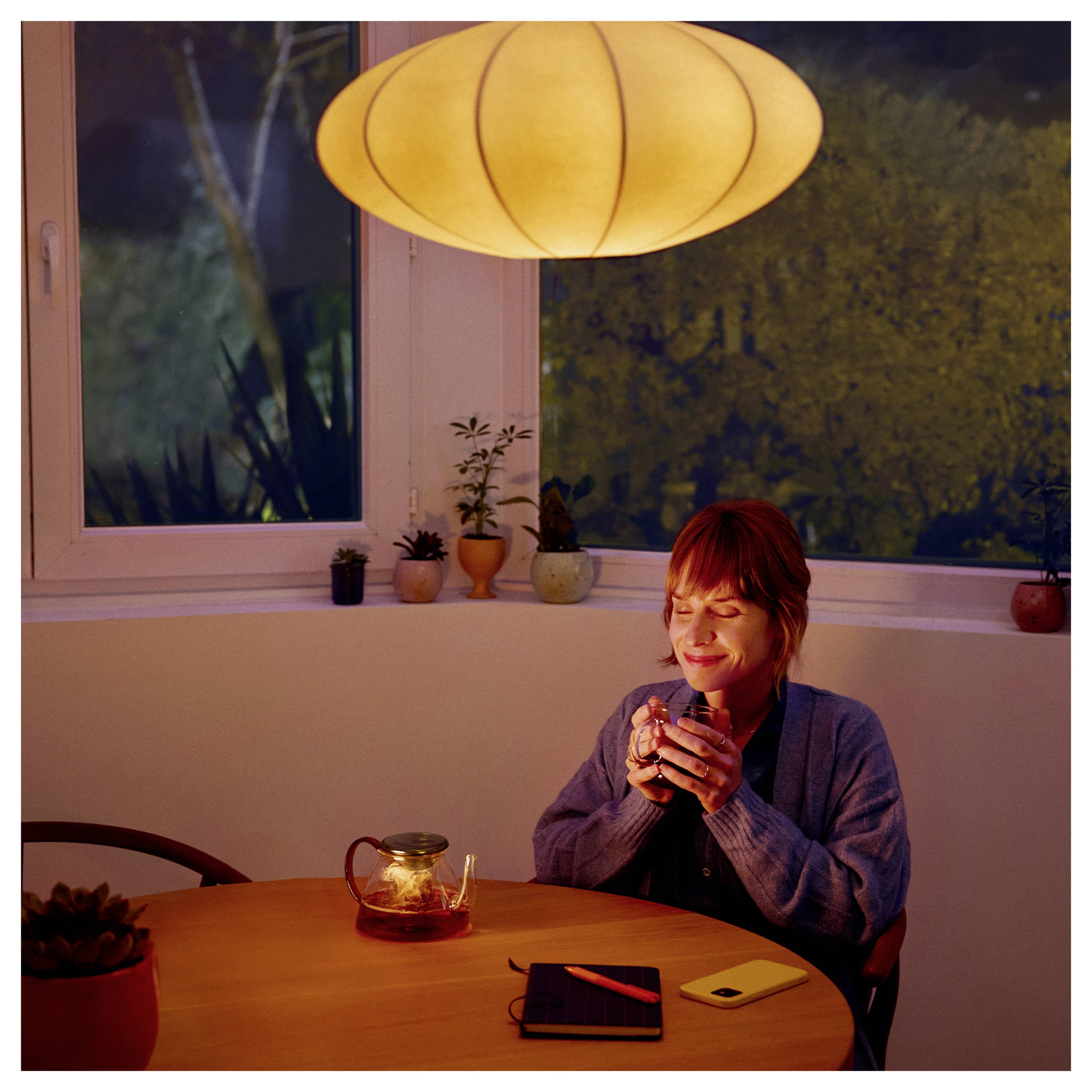 Osoba siedząca przy stole nocą, trzymająca kubek i uśmiechająca się. Na stole pod ciepło świecącą lampą stoją dzbanek do herbaty i notes.