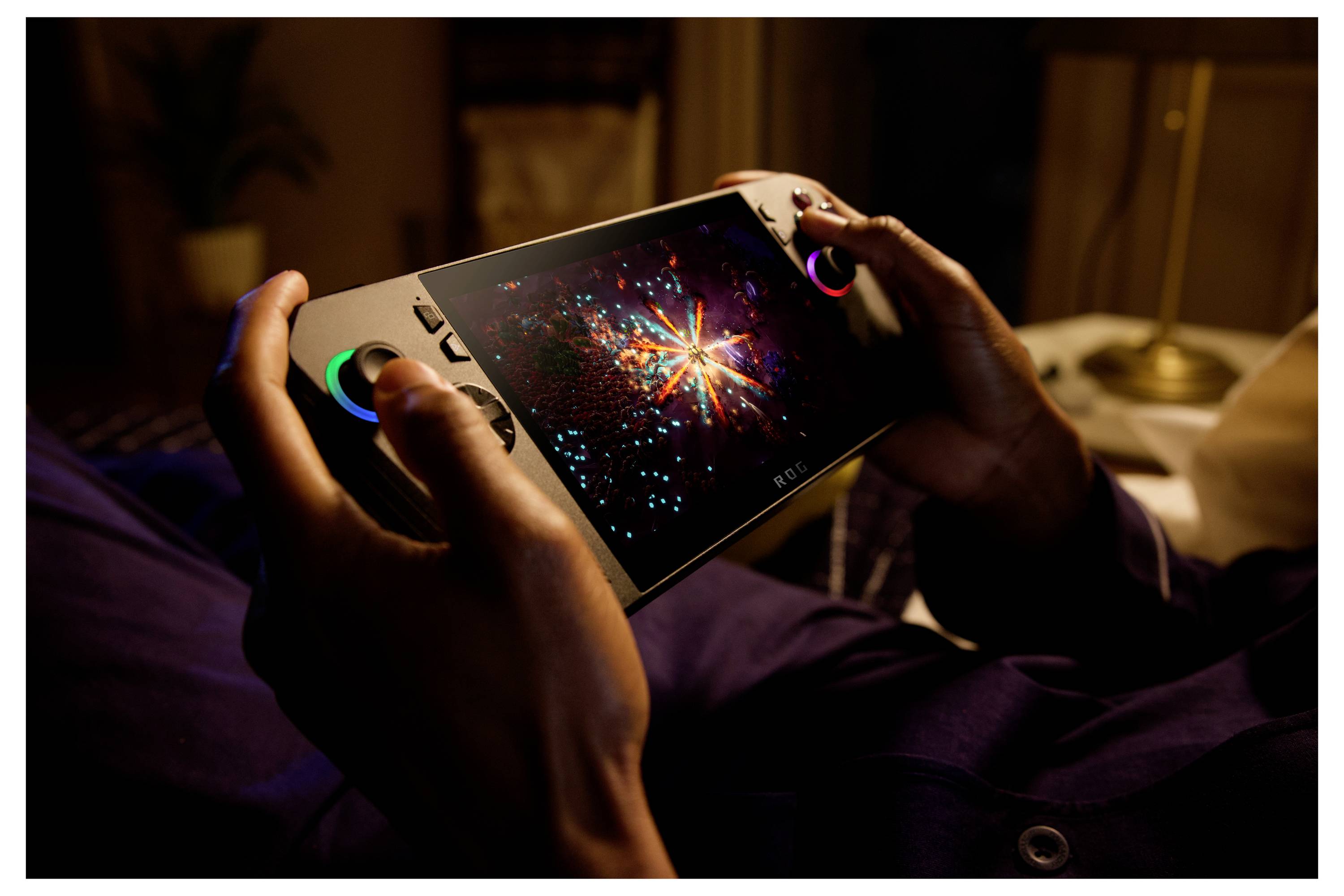 Osoba trzyma przenośną konsolę do gier, na której ekranie wyświetla się kolorowy wzór wybuchu, w słabo oświetlonym pomieszczeniu.