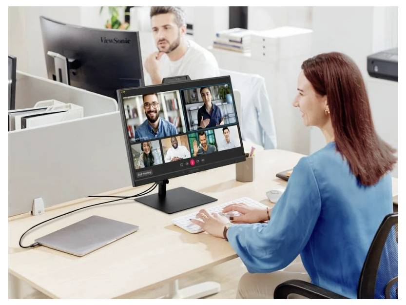 Kobieta siedząca przy biurku bierze udział w wideokonferencji na ekranie swojego komputera, na którym wyświetlonych jest wielu uczestników. Obok leży otwarty laptop.